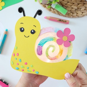 Könnte beinhalten: Ein gelbes Schnecken-Bastelprojekt aus Papier mit einem spiralförmigen, regenbogenfarbenen Gehäuse, schwarzen Pfeifenreiniger-Fühlern und kleinen bunten Punkten auf dem Körper. Eine rosa Papierblume ist am Gehäuse befestigt und das Gesicht ist gezeichnet.