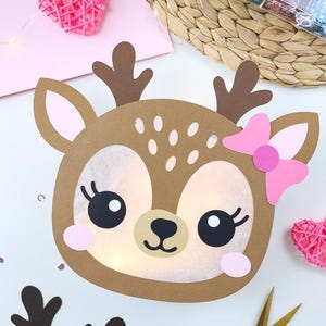 May include: A brown paper deer-shaped light with a pink bow, large black eyes with eyelashes, and white spots on the forehead. The face features a translucent centre illuminated by internal lights, accented by pink cheeks and dark brown antlers.