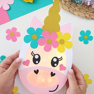 Könnte beinhalten: Ein rosa-weißes Einhorn-Bastelobjekt mit einer goldenen Glitzer-Hupe und bunten Blumenakzenten. Das Einhorn hat große Augen, herzförmige Wangen und eine leuchtende Mitte. Zusätzliche Blumenausschnitte sind verstreut.