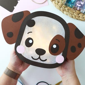 Könnte beinhalten: Eine handgefertigte Papierlaterne in Hundeform mit einem durchscheinenden weißen Gesicht und dunkelbraunem Rand, braunen Ohren mit dunklen Punkten, schwarzen Augen, einer schwarzen Nase und Mund sowie zwei rosa Wangen, beleuchtet durch warmes Licht.