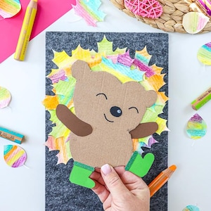 May include: A handmade craft project featuring a smiling bear made of cardboard, surrounded by colorful leaves. The bear has brown arms and a green boots. The background is a gray felt board. Crayons and leaf cutouts are scattered around.