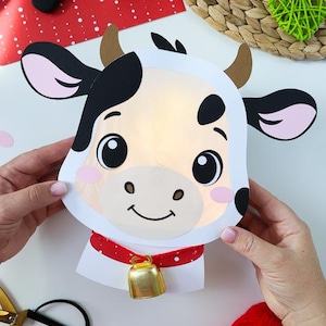 May include: A handmade cow-shaped paper lantern with a smiling face, black spots, and pink ears. It has a red collar with a gold bell. The lantern is lit from within, creating a warm glow. A pair of scissors and a ball of red yarn are nearby.