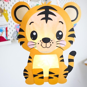Puede incluir: Un farol de papel colgante con forma de tigre hecho de cartulina naranja y amarilla, con rayas negras y una cuerda blanca. La cara del tigre presenta grandes ojos negros, una pequeña nariz negra y mejillas rosadas con luz interior.