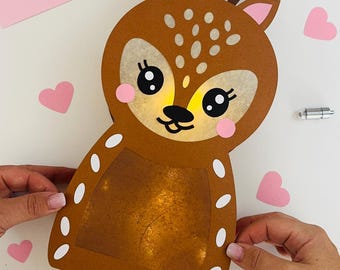 May include: A brown paper deer craft with white polka dots, pink ears, and black eyes. The deer has a light inside and is lit up. The deer is on a white surface with pink hearts and a pair of gold scissors.