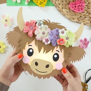 Puede incluir: Una cabeza de vaca Highland de papel con pelaje marrón claro, dos cuernos beige y una corona de flores artificiales multicolores. La cara tiene grandes ojos negros, un hocico beige y mejillas rosas. El diseño está iluminado desde atrás.