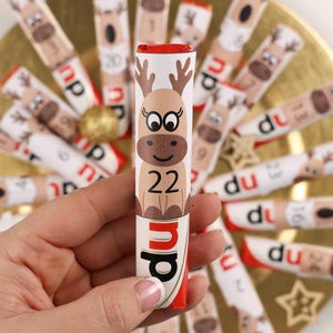 Könnte beinhalten: Ein Schokoladen-Adventskalender mit einem Rentier-Design. Jede Tages-Schokolade ist in einem kleinen Papier-Röhrchen mit einer Nummer und einer Rentier-Illustration verpackt. Der Kalender ist in einem Kreis auf einem goldenen Teller angeordnet.