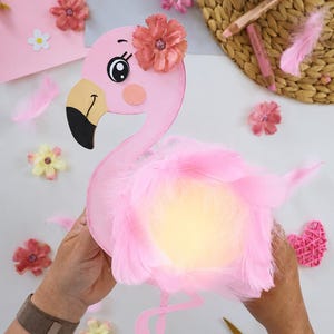 May include: A pink flamingo craft cutout featuring a large black and beige beak, a decorative peach-coloured fabric flower on its head, and a fluffy body made of light pink feathers. The flamingo has a cartoon-style eye with long eyelashes.