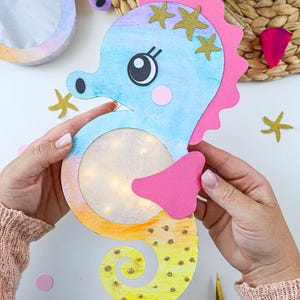 May include: A seahorse-shaped craft made of paper with a blue, pink, and yellow gradient finish. It features a circular cutout in the centre containing small glowing lights, pink fins, a pink dorsal ridge, and gold glitter star accents.