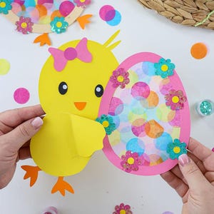 Könnte beinhalten: Ein gelbes Küken mit einer rosa Schleife hält ein rosa Ei mit einem bunten Punkte-Muster und Glitzerblumen. Das Ei ist geöffnet, um einen bunten Seidenpapier-Hintergrund zu zeigen.