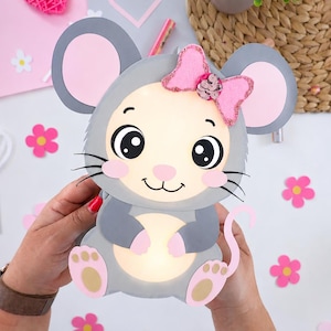 Puede incluir: Una lámpara de papel en capas con forma de ratón, cuerpo gris, vientre color crema iluminado, orejas grandes con centros rosas y un lazo rosa brillante con lentejuelas en la cabeza. El ratón tiene ojos negros, nariz rosa y almohadillas rosas.