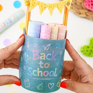 May include: A cylindrical blue pencil holder featuring the text Back to School in colorful chalk-style lettering. The holder is decorated with small icons like a globe, apple, and heart. Two yellow pencils support a yellow triangular banner.
