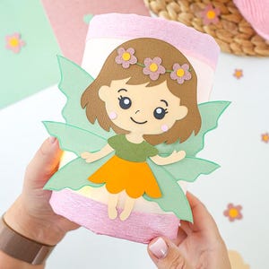 Könnte beinhalten: Eine zylindrische Leuchte, umwickelt mit hellrosa Krepppapier, mit einem Papieraufleger eines braunhaarigen Charakters mit grünen Flügeln, einem grünen Oberteil und einem orangefarbenen Rock. Der Charakter trägt einen Kranz aus drei kleinen Blüten.