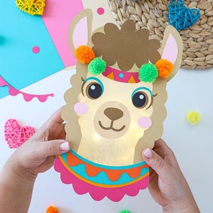 Puede incluir: Una lámpara troquelada con forma de llama que presenta una cara bronceada, pelo marrón y mejillas rosadas. La llama lleva una diadema colorida con pompones naranjas y verdes y un collar rosa y naranja brillante. Luces LED cálidas iluminan la cara.