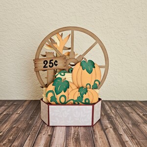 May include: A papercraft pumpkin stand with a wooden wheel, three pumpkins, and a sign that reads "25c".