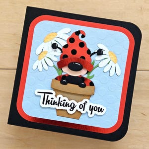 May include: A square greeting card with a black border, a red frame, and a light blue background with a subtle embossed dot pattern. The center features a ladybug-themed gnome in a tan flower pot, flanked by two white daisies, with the text Thinking of you.