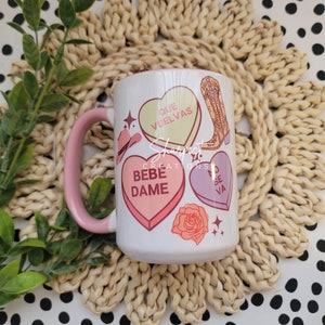May include: White ceramic mug with a pink handle. The mug features a colorful design with hearts, cowboy boots, and a rose. The hearts have Spanish text: "Que Vuelvas", "Bebe Dame", and "No Se Va".