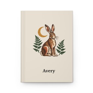 May include: A cream-colored hardcover journal featuring a central illustration of a brown rabbit sitting between two green fern fronds under a golden crescent moon. The name Avery is printed in black serif font below the illustration.