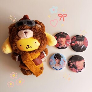 May include: A plush toy of a yellow character wearing a brown aviator hat, scarf, and jacket, with four round pinback buttons featuring images of a person. The toy has a brown fur-like mane.