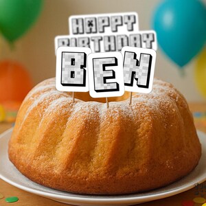 May include: A golden-brown bundt cake dusted with powdered sugar, topped with a "Happy Birthday BEN" cake topper. Colorful balloons and confetti decorate the background, creating a festive birthday scene.