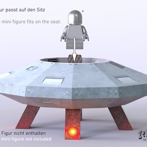May include: A gray papercraft model of a flying saucer with red legs and a small gray figure standing on the top. The text "Spielfigur passt auf den Sitz" and "Famous mini figure fits on the seat." is printed above the saucer. The text "* Figur nicht enthalten" and "mini figure not included" is printed below the saucer. The text "getide let's make it" is printed on the right side of the saucer.