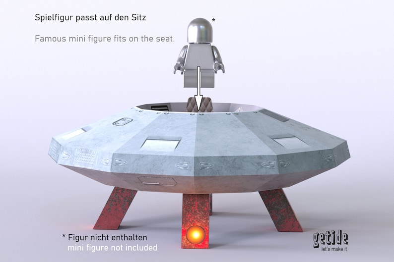 May include: A gray papercraft model of a flying saucer with red legs and a small gray figure standing on the top. The text "Spielfigur passt auf den Sitz" and "Famous mini figure fits on the seat." is printed above the saucer. The text "* Figur nicht enthalten" and "mini figure not included" is printed below the saucer. The text "getide let's make it" is printed on the right side of the saucer.