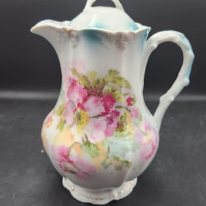 May include: A white porcelain creamer with a floral design. The creamer has a lid with a blue rim and a handle. The flowers are pink, yellow, and green.