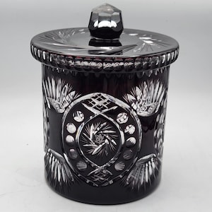 Puede incluir: Un tarro de cristal negro con tapa, que presenta intrincados diseños de vidrio cortado. El tarro tiene una forma redonda y un acabado liso y pulido.