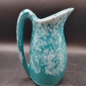 May include: A teal ceramic pitcher with a white speckled glaze. The pitcher has a rounded body and a curved handle.