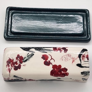 May include: A rectangular ceramic trinket box with a dark teal lid and a cream base. The base is decorated with a floral pattern in red and black. The box is open, showing the lid. Measurements are in centimetres.