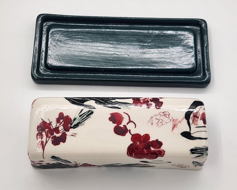 May include: A rectangular ceramic trinket box with a dark teal lid and a cream base. The base is decorated with a floral pattern in red and black. The box is open, showing the lid. Measurements are in centimetres.