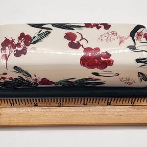 May include: Cream-coloured ceramic butter dish with a floral design in red and black on the lid. The base is dark green. The dish is approximately 15 cm long.