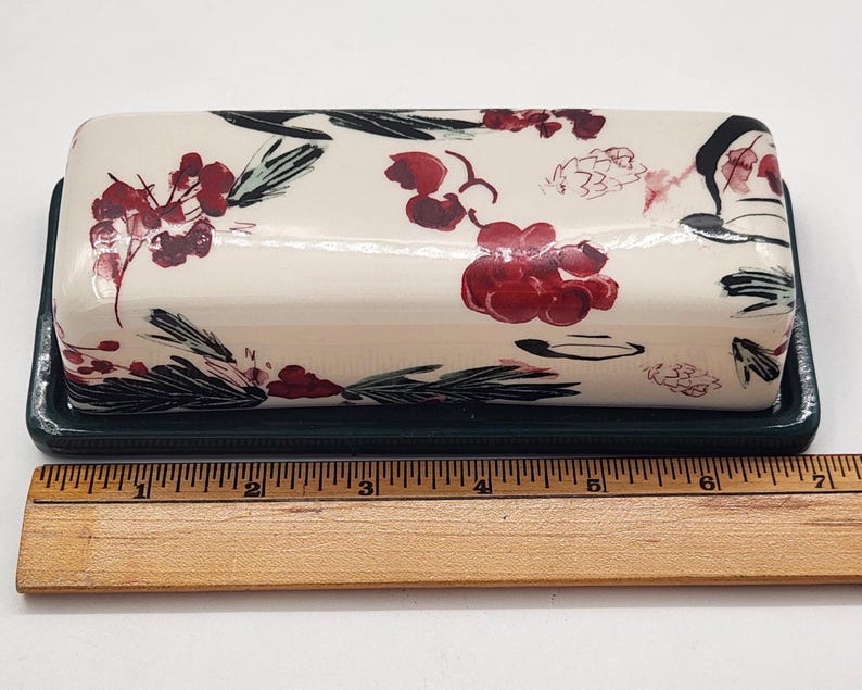 May include: Cream-coloured ceramic butter dish with a floral design in red and black on the lid. The base is dark green. The dish is approximately 15 cm long.