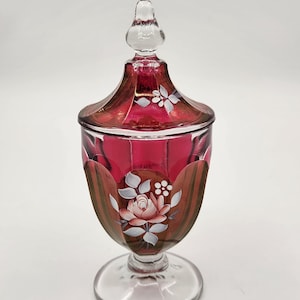 May include: A ruby red glass jar with a lid, featuring hand-painted floral designs. The jar has a clear glass base and a decorative finial on the lid. The floral design includes a pink rose and white accents.
