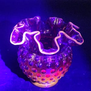 May include: A deep purple glass bowl featuring a hobnail texture across its exterior surface. The bowl has a wide, ruffled, and crimped rim that glows with a bright, light-colored edge under ultraviolet light, highlighting the intricate glasswork.