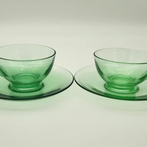 Op de afbeelding: Twee bijpassende groene glazen theekopjes en schotels. De kopjes hebben een klassiek ontwerp met een oor en staan op ronde schotels. Het glas is transparant en de set wordt gepresenteerd tegen een witte achtergrond.