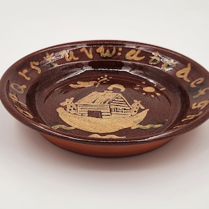 May include: A brown ceramic plate with a decorative design. The plate features a central image of a house on a boat, surrounded by an alphabet. The rim of the plate is a darker brown, and the base is a lighter shade of brown.