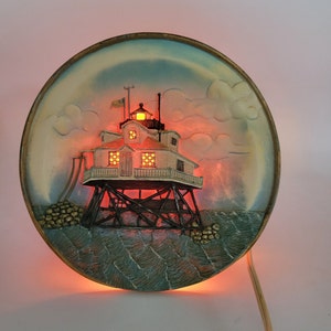 Puede incluir: Un plato de cerámica con una imagen 3D de un faro sobre pilotes en el océano. El faro está iluminado con un cálido resplandor naranja. El plato es redondo y tiene un borde marrón claro.