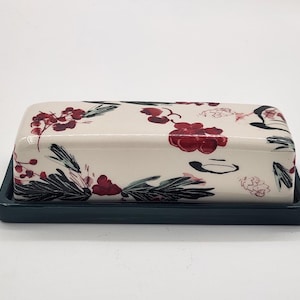 May include: A rectangular ceramic butter dish with a lid, decorated with a cream-coloured floral pattern in red, black, and green. The base is a solid dark teal colour, providing a contrasting backdrop.