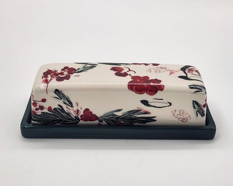 May include: A rectangular ceramic butter dish with a lid, decorated with a cream-coloured floral pattern in red, black, and green. The base is a solid dark teal colour, providing a contrasting backdrop.