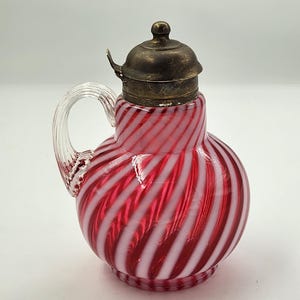 May include: A small glass pitcher featuring a red and white spiral striped pattern. The vessel has a clear glass handle and a tarnished metallic lid with a small knob on top. The body of the pitcher is rounded with a narrow neck and a ribbed base.