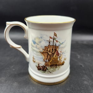 Pode incluir: Caneca de cerâmica branca com borda e alça douradas. A caneca apresenta uma ilustração detalhada de um navio alto com velas marrons e bandeiras coloridas. O fundo mostra um céu azul com nuvens. Uma peça decorativa.