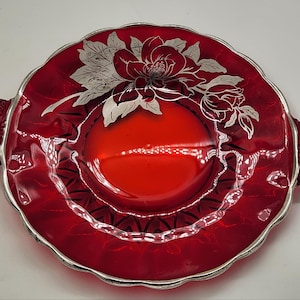 May include: A round, ruby red glass serving plate with two ornate handles. The plate features a scalloped edge trimmed in silver and a prominent silver floral design of a large blossom and leaves centered on the upper portion of the glass surface.