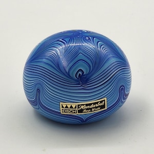 Op de afbeelding: Blauwe glazen presse-papier met een wervelend patroon van licht- en donkerblauwe lijnen. De presse-papier heeft een afgeronde vorm en een klein rechthoekig etiket met de tekst "Eisch Handarbeit Bayern Wald".