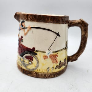 May include: A ceramic pitcher with a brown handle and a whimsical scene of a man in a red car fishing with a long pole. The scene is painted in bright colors and includes a brown bear and a rabbit. The pitcher has the word "HUNG" written on the bottom.