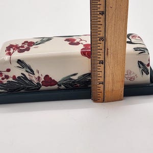 May include: A rectangular ceramic butter dish with a lid, on a dark green base. The cream-coloured lid is decorated with red floral and green leaf designs. The dish is approximately 10 cm long.