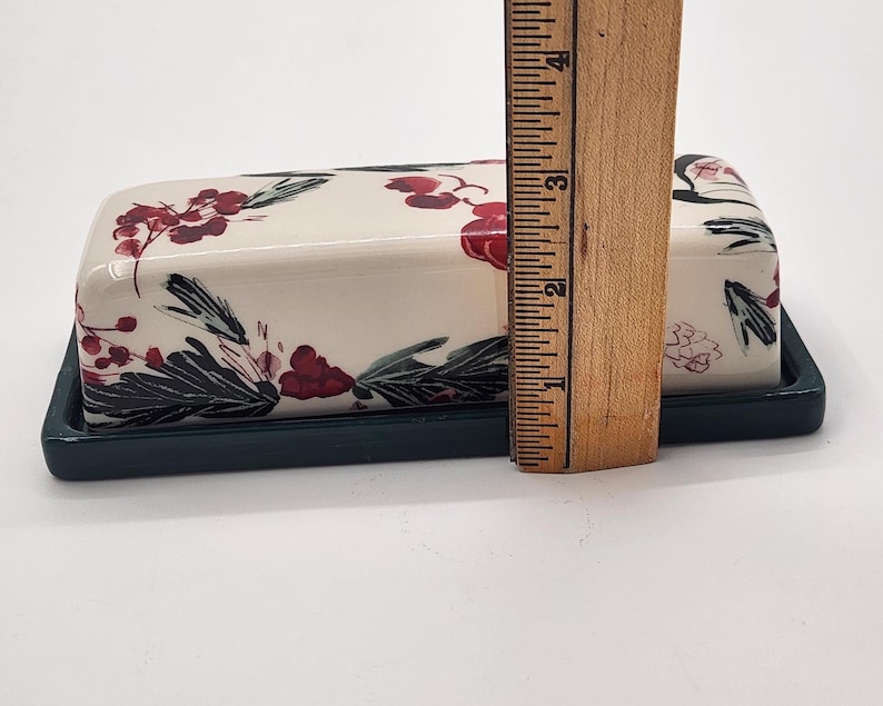 May include: A rectangular ceramic butter dish with a lid, on a dark green base. The cream-coloured lid is decorated with red floral and green leaf designs. The dish is approximately 10 cm long.