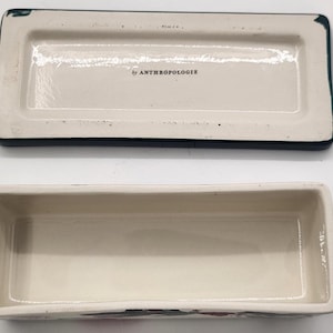 May include: A rectangular ceramic trinket box with a lid, featuring a cream-coloured interior and a dark blue border. The lid is off, revealing the box's interior. The text "by ANTHROPOLOGIE" is visible on the lid.