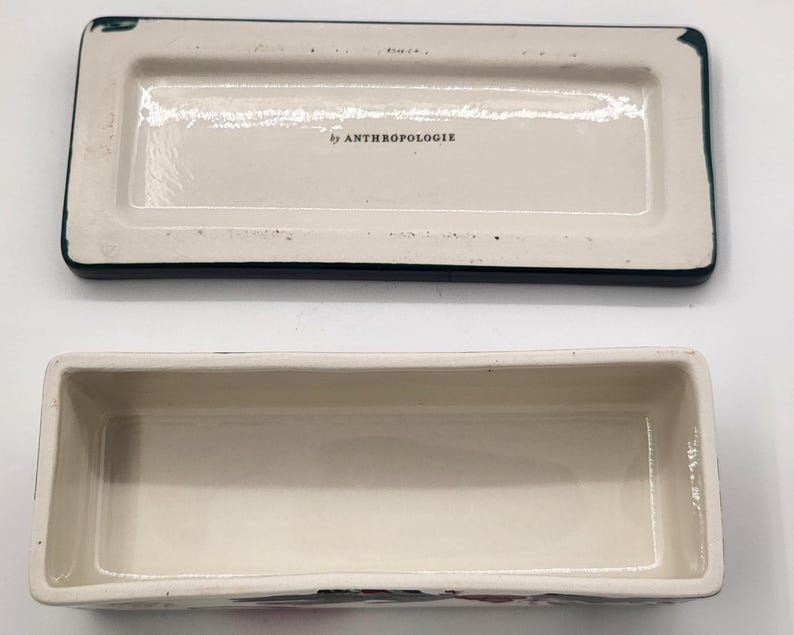 May include: A rectangular ceramic trinket box with a lid, featuring a cream-coloured interior and a dark blue border. The lid is off, revealing the box's interior. The text "by ANTHROPOLOGIE" is visible on the lid.