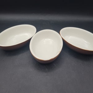 May include: Three white oval ceramic ramekins with brown rims. The ramekins are arranged in a row on a black surface.