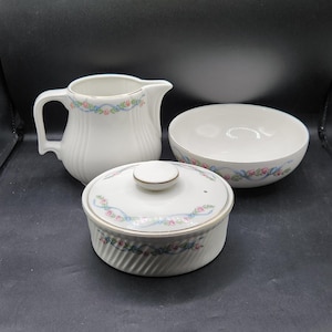 Könnte beinhalten: Eine weiße Keramik-Milchkanne, eine Zuckerdose mit Deckel und eine Schüssel mit einem Blumendesign mit rosa Rosen und blauen Bändern. Das Set ist im Vintage-Stil.